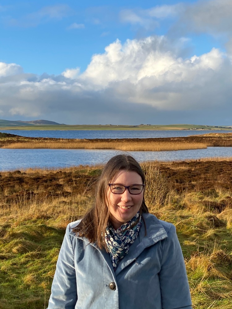 A colour photo of Ellen, a white woman with brown hair and glasses wearing a blue coat and scarf. She is stood in front of the Orkney seascape.